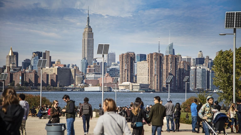 People on a promenade with solar panels and buildings in background. View of Manhattan is against sky.