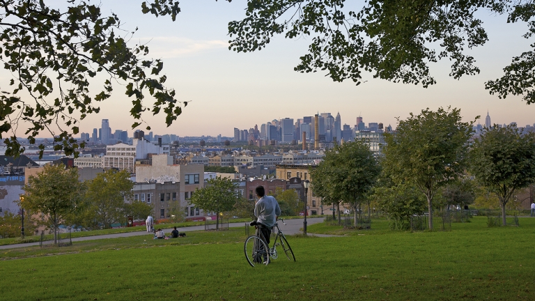 A person with a bicycle stands on a grassy hill in Sunset Park, overlooking a New York City skyline under a clear sky. Trees frame the scene.