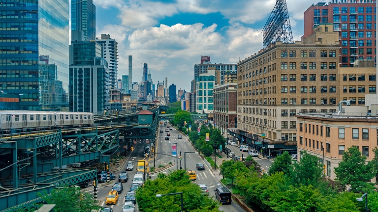 A busy urban street with cars and yellow cabs runs between tall buildings. A subway train travels on an elevated track to the left. The city skyline is visible in the background under a partly cloudy sky.