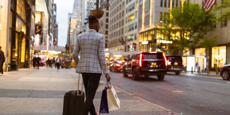 Person in plaid jacket walking on New York City sidewalk with a rolling suitcase and shopping bags. Busy Fifth Avenue and tall buildings in the background.